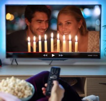 Hand holding TV remote pointed at TV featuring couple looking at Hanukkah menorah